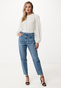 Blusa bianca a maniche lunghe con ricamo testurizzato, abbinata a jeans azzurri a vita alta e sandali neri con tacco. Outfit semplice e casual.
