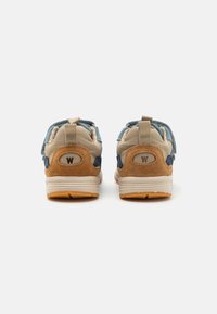 Bakvy av sneakers med en beige ovandel, blå och tan mockadetaljer, vit mellansula och gummisula. Har en ögla och "W"-logotyp.