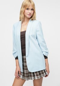 Svetlomodrý blejzer s mierne vypchatými rukávmi, otvorený dizajn, skombinovaný s hnedým topom a kockovanou mini sukňou s skladaným lemom.