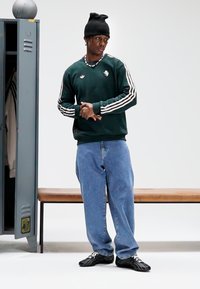 Sweatshirt verde de manga comprida com riscas brancas e logótipo, combinada com jeans largos azul claro e sapatilhas desportivas pretas, em pé ao lado de um cacifo.