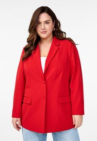 Roter Blazer mit strukturiertem Design, Ein-Knopf-Verschluss und zwei Fronttaschen. Kombiniert mit hellblauen Jeans.