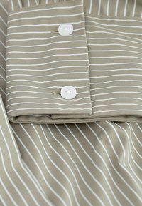Beige stof met witte strepen, met een opgevouwen kraag en twee witte knopen. Gladde textuur met een frisse uitstraling.