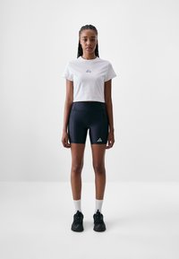 White cropped t-shirt with small blue logo, paired with black fitted shorts featuring a triangle logo. Black sneakers and white socks complete the look.