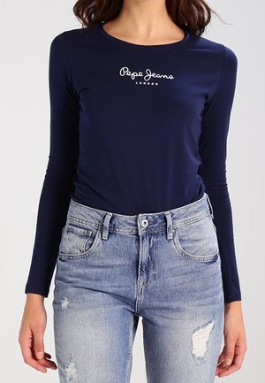 Kobieta ubrana w granatową koszulkę z długim rękawem z logo "Pepe Jeans London" oraz jasnoniebieskie, przetarte dżinsy z przednimi kieszeniami i szlufkami na pasek.