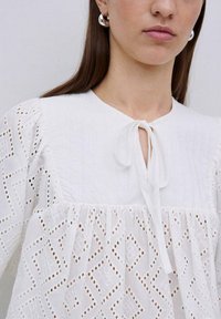 Blusa bianca con scollatura rotonda e chiusura con lacci, maniche a sbuffo e sofisticata ricamo ad occhio in una trama a diamante.
