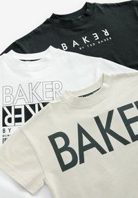 Tres camisetas exhibidas: negra, blanca y beige claro. Cada una presenta el texto en negrita "BAKER" en diferentes estilos y tamaños. Material de algodón suave.