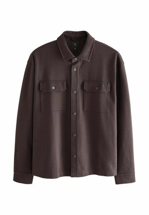Hombre con chaqueta marrón oscuro abotonada sobre sudadera con capucha beige, pantalones negros y zapatos negros, de pie frente a un fondo gris liso.