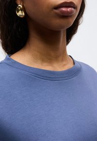 Κοντινό πλάνο του λαιμού και των χειλιών ενός ατόμου που φοράει μπλε μπλούζα με στρογγυλή λαιμόκοψη και ένα μεγάλο λαμπερό χρυσό σκουλαρίκι στο αριστερό αυτί.