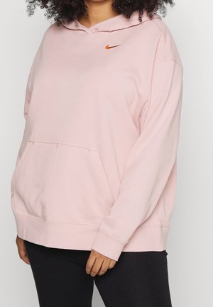 Osoba nosząca za dużą jasnoróżową bluzę Nike z pomarańczowym logo Swoosh i czarne spodnie, stojąca na jednolitym tle.