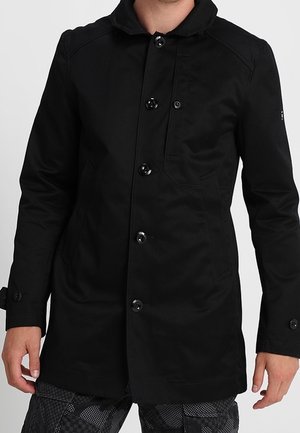 Homme portant un long manteau noir boutonné avec col et un pantalon sombre à motifs sur un fond clair uni.