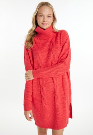 Jeune femme aux longs cheveux blonds portant une robe-pull à col roulé côtelée corail rose texturée, souriant légèrement avec les bras croisés.
