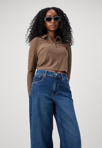 Smeđi rebrasti longsleeve top s ovratnikom i patentnim zatvaračem, uparen s visokim plavim širokim trapericama. Model nosi tirkizne pravokutne sunčane naočale.