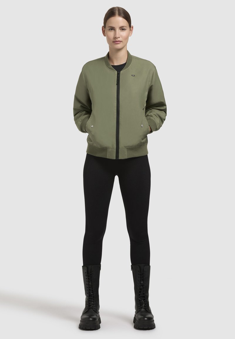 Olivgrüne Bomberjacke mit Reißverschluss, gerippten Bündchen und Saum; getragen über schwarzen Leggings und schwarzen Schnürboots.