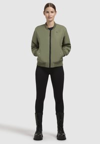 Olijfgroene bomberjack met een ritssluiting, geribde manchetten en decoratieve drukknoopjes, gedragen met zwarte leggings en legerlaarzen.