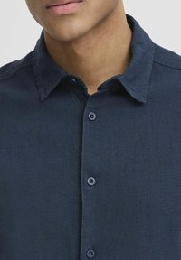 Camisa azul marino de botón en un tejido texturizado; cuenta con un collar clásico, siete botones en la parte delantera y un aspecto limpio y a medida.