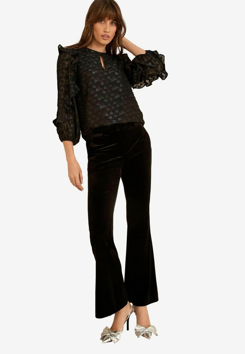 Blusa nera con texture e maniche a volant, caratterizzata da uno scollo a chiave. Abbinata a pantaloni neri in velluto a zampa d'elefante e tacchi adornati da fiocchi argentati.