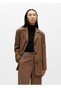 Bruine op maat gemaakte blazer met een gestructureerd ontwerp, zwarte coltrui en bijpassende broek. Voorzien van knoopsluiting en zijzakken.