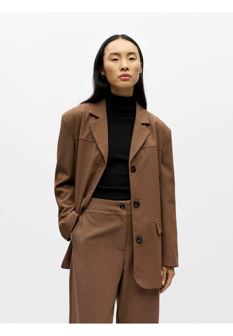 Bruine op maat gemaakte blazer met een gestructureerd ontwerp, zwarte coltrui en bijpassende broek. Voorzien van knoopsluiting en zijzakken.