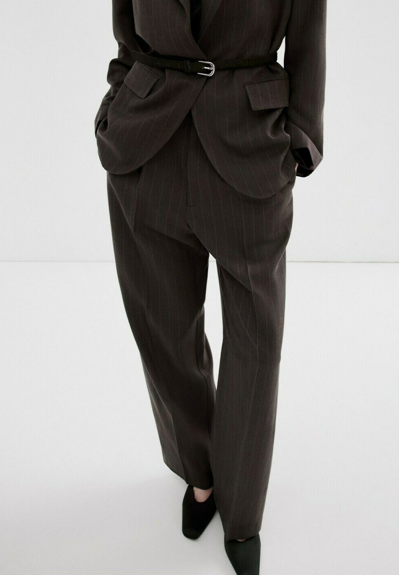 Costum întunecat cu dungi subțiri, format dintr-un sacou cu curea și pantaloni largi și lejeri, purtat cu pantofi negri cu vârf ascuțit pe fundal alb.