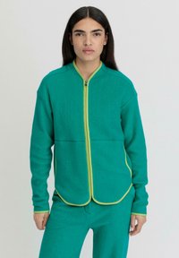 Giacca in pile verde con colletto alto, zip gialla e dettagli a contrasto. Il design presenta un orlo arrotondato e maniche lunghe per una vestibilità comoda.