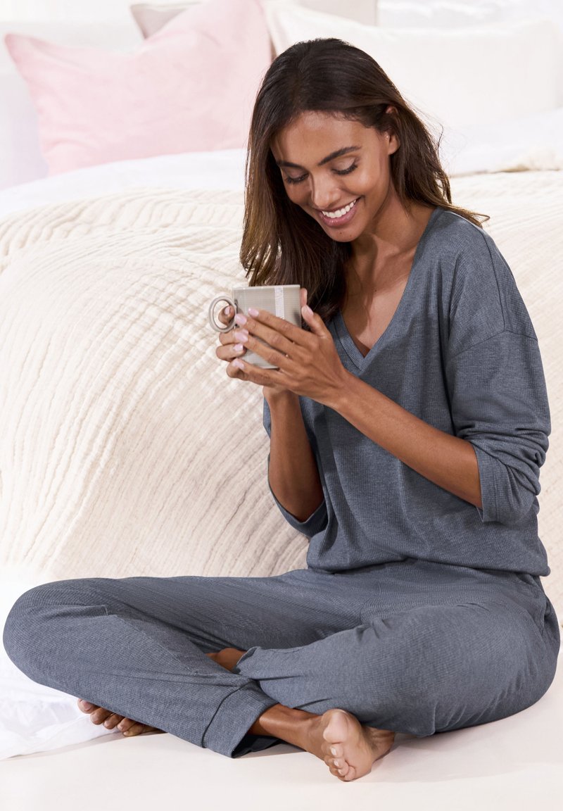 Gray lounge set with a V-neck top and tapered pants, made of soft material. Individual sits cross-legged, holding a mug, on a textured blanket.