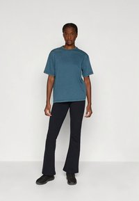 Turkizno modra bombažna majica z okroglim izrezom, kombinirana s črnimi razširjenimi hlačami in črnimi čevlji, stoji pred nevtralnim ozadjem.