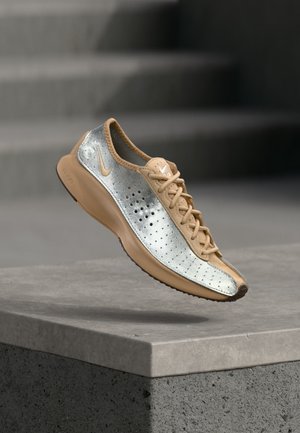 Baskets Nike tan et argent avec perforations et lacets, flottant au-dessus d'un rebord en béton sur un fond gris flou.