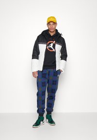 Chaqueta puff en blanco y negro, sudadera con capucha y logo, pantalones de chándal a cuadros en azul y verde, combinados con zapatillas de deporte verdes de caña alta.