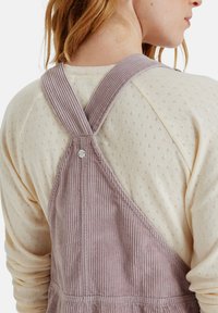 Lichtpaarse corduroy tuinbroek met brede gestreepte bandjes en een zilveren knoop, gedragen over een crèmekleurig textuur langemouwen shirt.