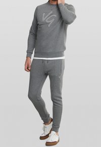 Felpa grigia con un logo "VG" testurizzato, abbinata a pantaloni jogger grigi coordinati. Presenta polsini a coste e tasche laterali, indossata con sneakers bianche.