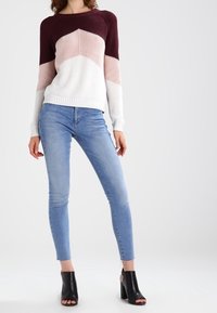 Eine Frau trägt einen farblich abgestimmten Pullover in Burgunderrot, Pink und Weiß, kombiniert mit hellblauen Skinny-Jeans und schwarzen offenen Absatzstiefeletten.