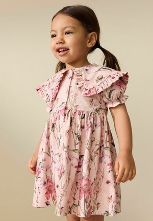 Ung pige med brunt hår i to fletninger iført en lyserød blomstret kjole med flæset ærmer, smilende og kigger let til siden.