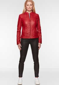 Rote Lederjacke mit Stehkragen, vorderem Reißverschluss und senkrechten Nahtdetails; getragen über schwarzen Skinny-Hosen und weißen Sneakers.