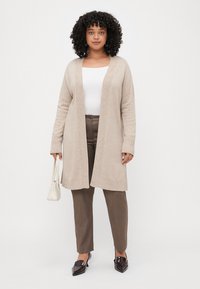 Cardigan en tricot beige avec des poignets et un ourlet côtelés, design ouvert à l'avant. Associé à un pantalon marron et des mules à talons foncés, tenant un sac blanc.