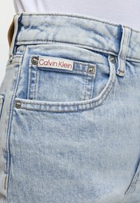 Poche de jean en denim bleu clair avec une étiquette blanche Calvin Klein cousue au-dessus de la poche et des rivets métalliques visibles.