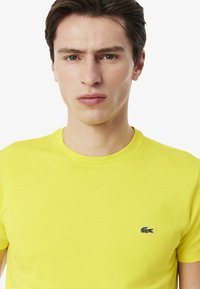 Felgekleurde gele t-shirt met korte mouwen en ronde hals, voorzien van een klein geborduurd krokodillenlogo op de linkerborst. Glad van textuur.