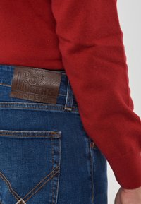 Jeans in denim blu con cuciture marroni e etichetta in pelle "Roy Roger's", indossati con una maglietta rossa a maniche lunghe.