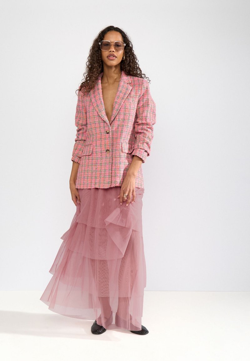 Femme portant un blazer rose à carreaux surdimensionné, une jupe en tulle rose à plusieurs couches, des lunettes de soleil et des chaussures tressées noires, debout sur fond blanc.