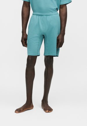 Hombre con pantalones cortos elásticos hasta la rodilla de color azul verdoso y camiseta a juego de manga corta, de pie descalzo sobre fondo blanco.