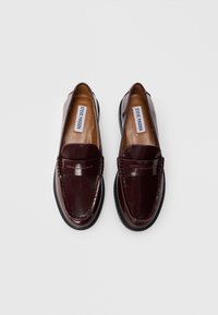 Une paire de mocassins penny en cuir marron foncé avec semelles noires et étiquettes « Steve Madden » à l’intérieur, présentée sur fond blanc.