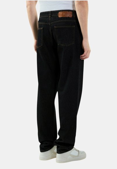 Persona che indossa jeans neri a vestibilità larga con tasche posteriori e patch di pelle marrone con logo GCDS, abbinati a sneakers bianche e una maglietta bianca.