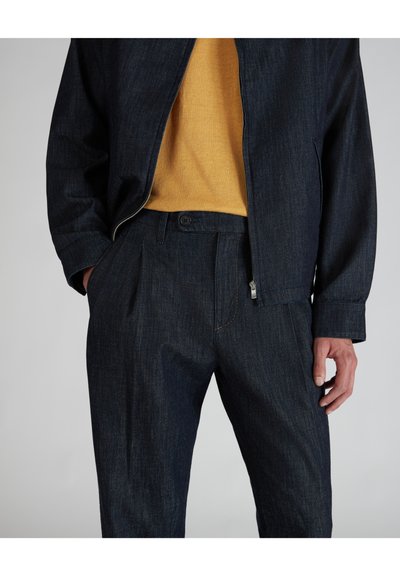 Giacca e pantaloni in denim scuro, con chiusura a zip, dettagli di cucitura arancione e una finitura testurizzata. L'abbinamento è completato da una maglietta gialla.