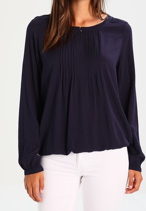 Navyblauwe blouse met lange mouwen, een ronde halslijn, subtiele plooien, gestructureerde stof en elastische boorden. Gecombineerd met witte broeken.