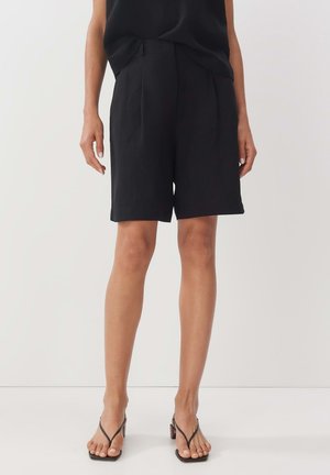 Schwarze Leinenkurzshorts mit maßgeschneiderter Passform, versehen mit Falten und einer Länge bis zur Mitte des Oberschenkels. Glatte Textur und seitliche Taschen. Kombiniert mit minimalistischem Schuhwerk.
