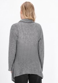 Donna con capelli biondi che indossa un lungo cardigan grigio dalla trama e pantaloni neri, vista da dietro su uno sfondo bianco semplice.