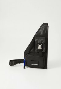 Musta olkalaukku, joka on valmistettu teksturoidusta nylonista. Siinä on vino muotoilu, säädettävä olkahihna, etutasku hopeisilla metalliosilla sekä sininen vetoketjun vetolaite.