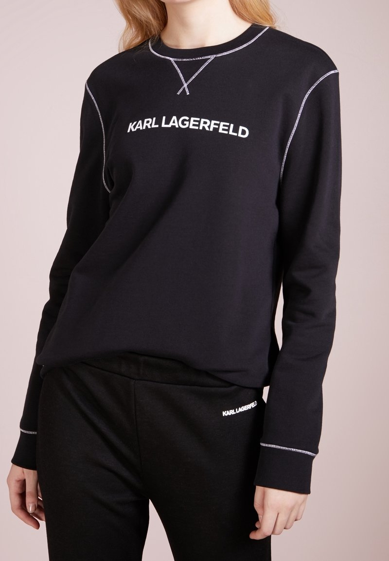 Schwarzes Sweatshirt- und Hosenset mit langen Ärmeln, weißen Kontrastnähten und "KARL LAGERFELD"-Logo auf der Brust und links an der Hüfte.