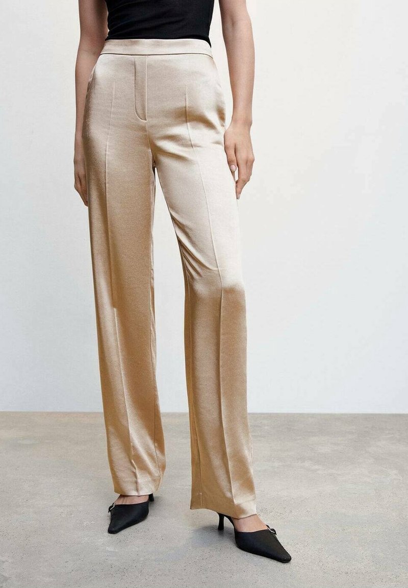 Femme portant un pantalon satin beige à jambes larges avec une taille haute, associé à des escarpins noirs à bout pointu et un haut noir sans manches.