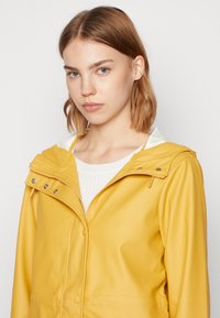 Jachetă galbenă impermeabilă cu un design ajustat, închidere cu fermoar, accesorii din metal argintiu și o glugă albă contrastantă. Textură netedă, fără modele vizibile.