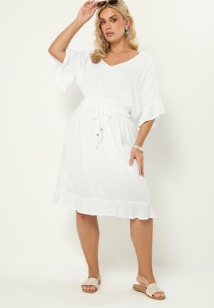 Blondynka w białej sukience do kolan z wiązanym w talii sznurkiem, trzymająca okulary przeciwsłoneczne na głowie, stojąca na jednolitym jasnym tle.